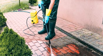 Man cleaning red, conrete pavement block using high pressure water cleaner. High pressure cleaning. Man wearing waders, protective, waterproof trousers and gloves doing spring jobs outdoors.