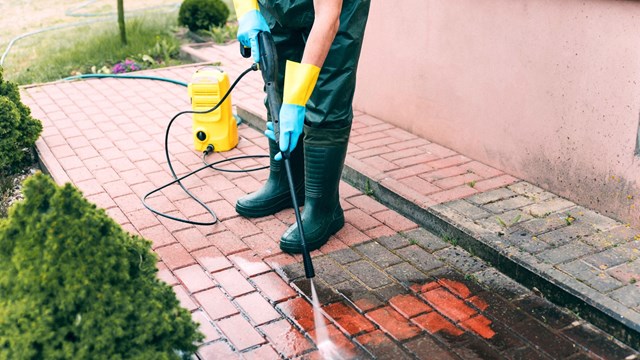 Man cleaning red, conrete pavement block using high pressure water cleaner. High pressure cleaning. Man wearing waders, protective, waterproof trousers and gloves doing spring jobs outdoors.