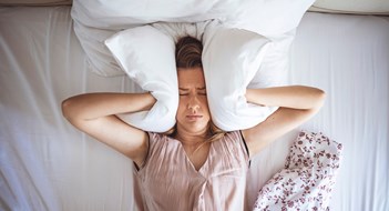 Young woman trying to sleep but disturbed by noisy neighbors and covering ears with pillows. I can's sleep with all that noise! Sleep problems. Angry woman disturbed with a noise trying to sleep