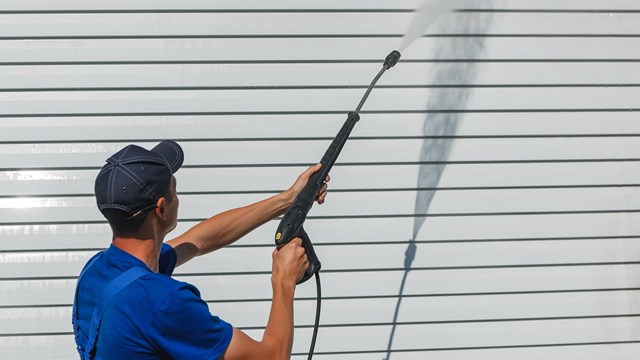 Worker in overalls washes a white wall from a siding with a water gun. Cleaning service for washing buildings and facades