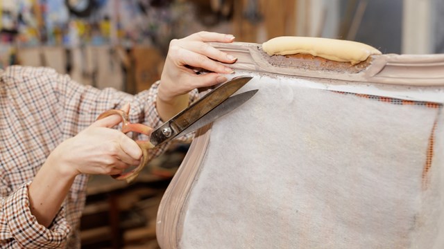 Close-up of hands cutting upholstery fabric with large scissors while restoring vintage furniture in a workshop.